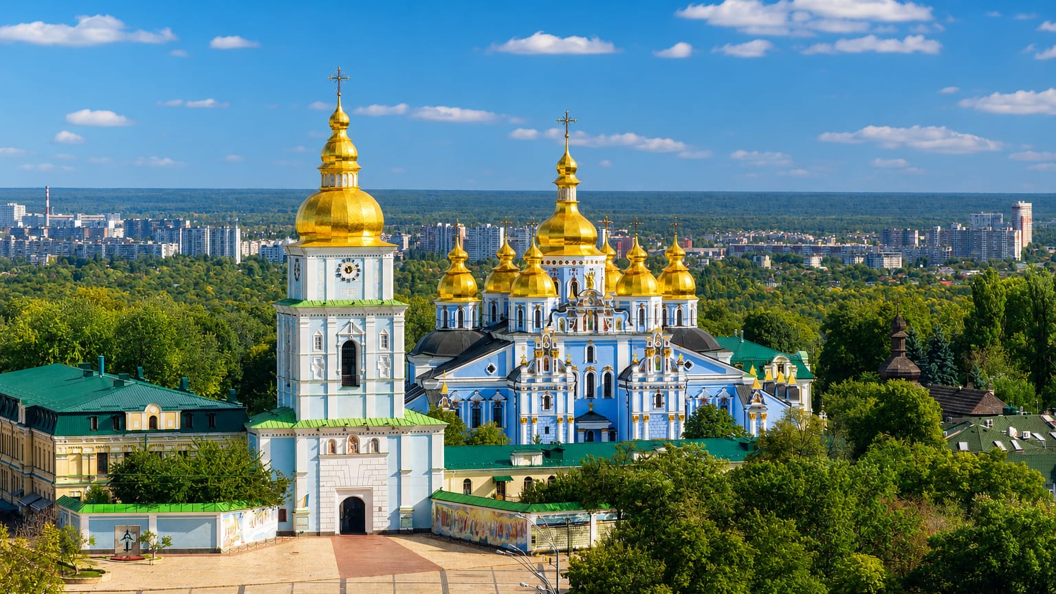 Kyiv's blue-and-gold St. Michael's Golden-Domed Monastery framed by green treetops under a clear sky.