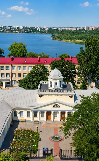 Riverside view of Mykolayiv, Ukraine, with a white colonnaded mansion and red-roofed building beside the wide estuary and a distant bridge.