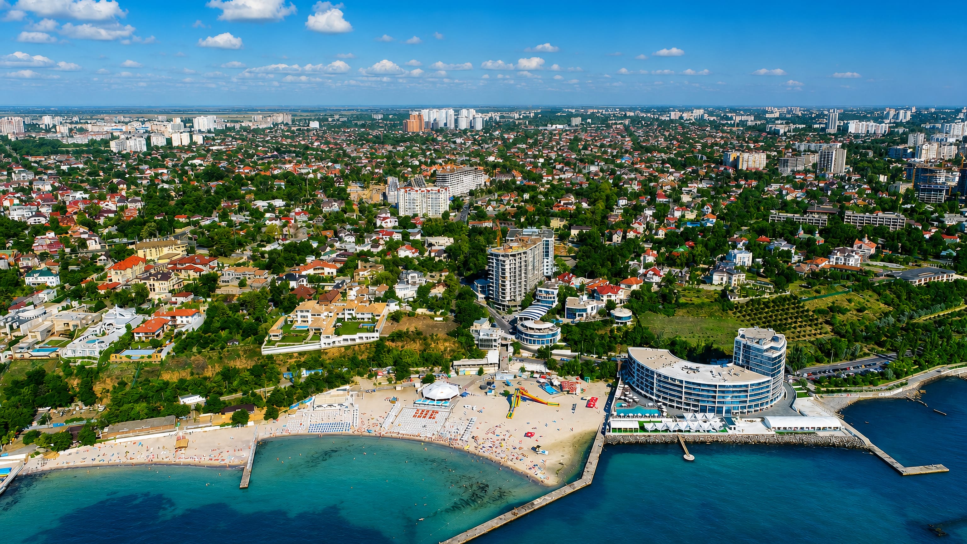 Aerial view of Odesa's Black Sea coastline with a curved beachfront resort, sandy beach and leafy suburbs.