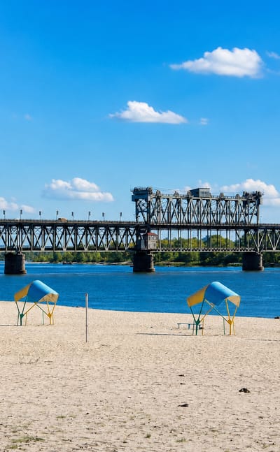 Sandy Dnieper riverbank at Kremenchuk, Ukraine, with beach shelters below the long steel Kriukiv railway bridge.