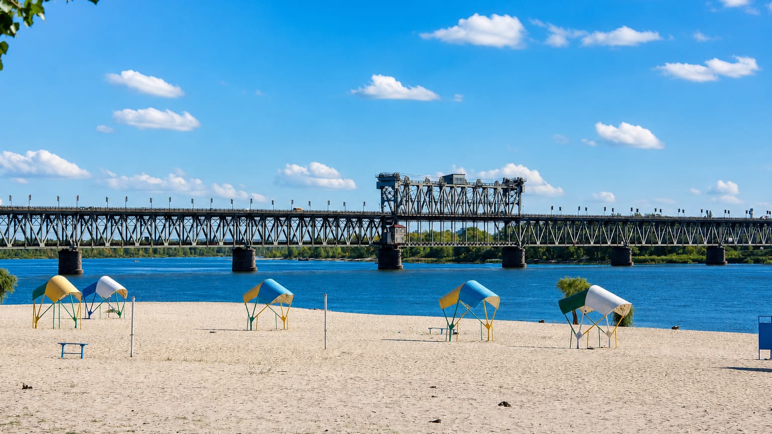 Sandy Dnieper riverbank at Kremenchuk, Ukraine, with beach shelters below the long steel Kriukiv railway bridge.