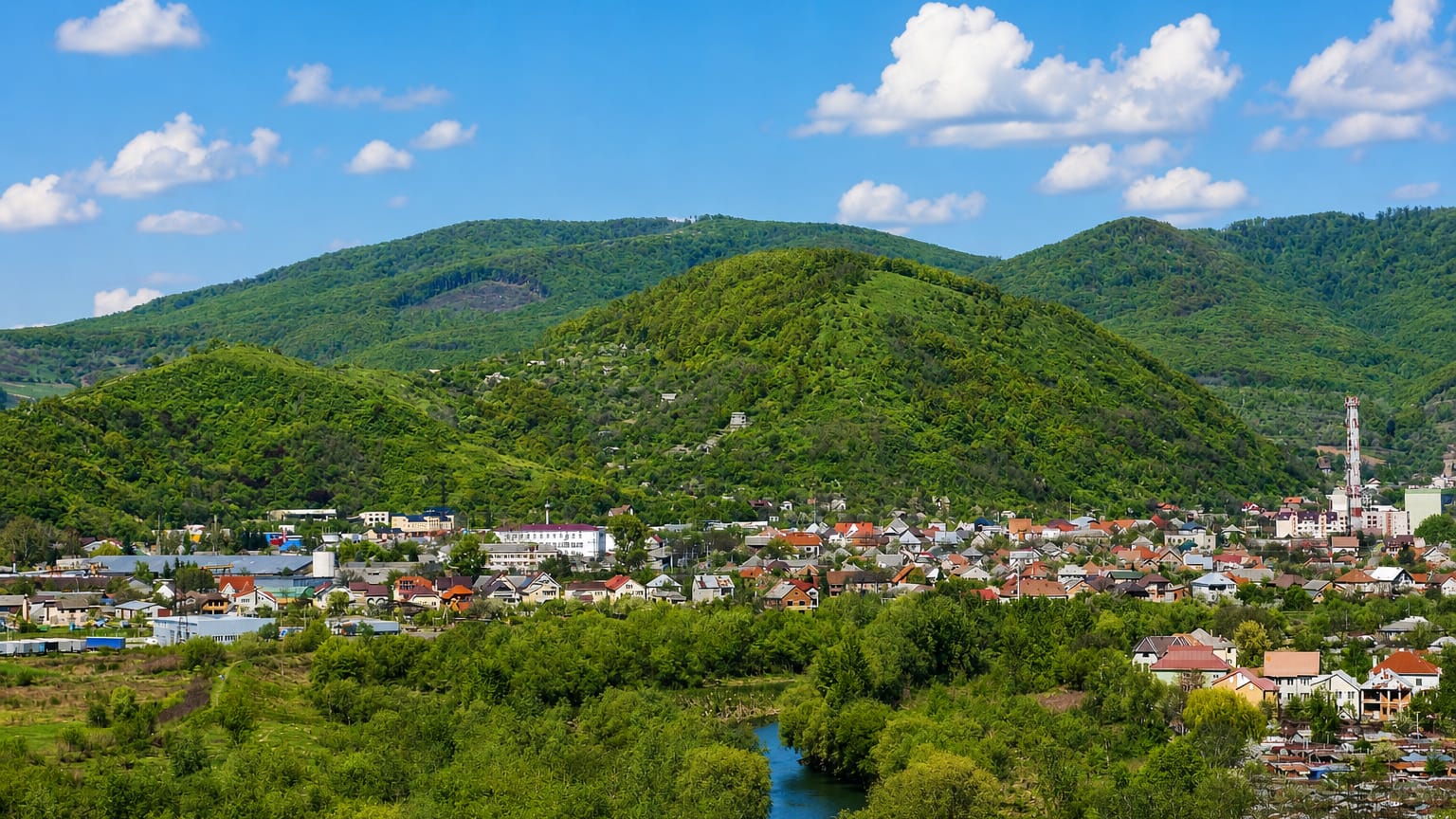 Mukachevo, Ukraine, set in a green Carpathian valley with a river winding past red-roofed houses below forested hills.