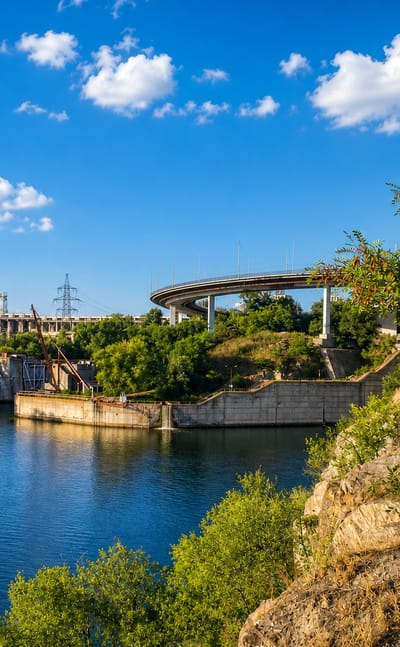 The Dnieper River at Zaporizhzhia with the DniproHES dam and a curving road bridge on rocky banks.