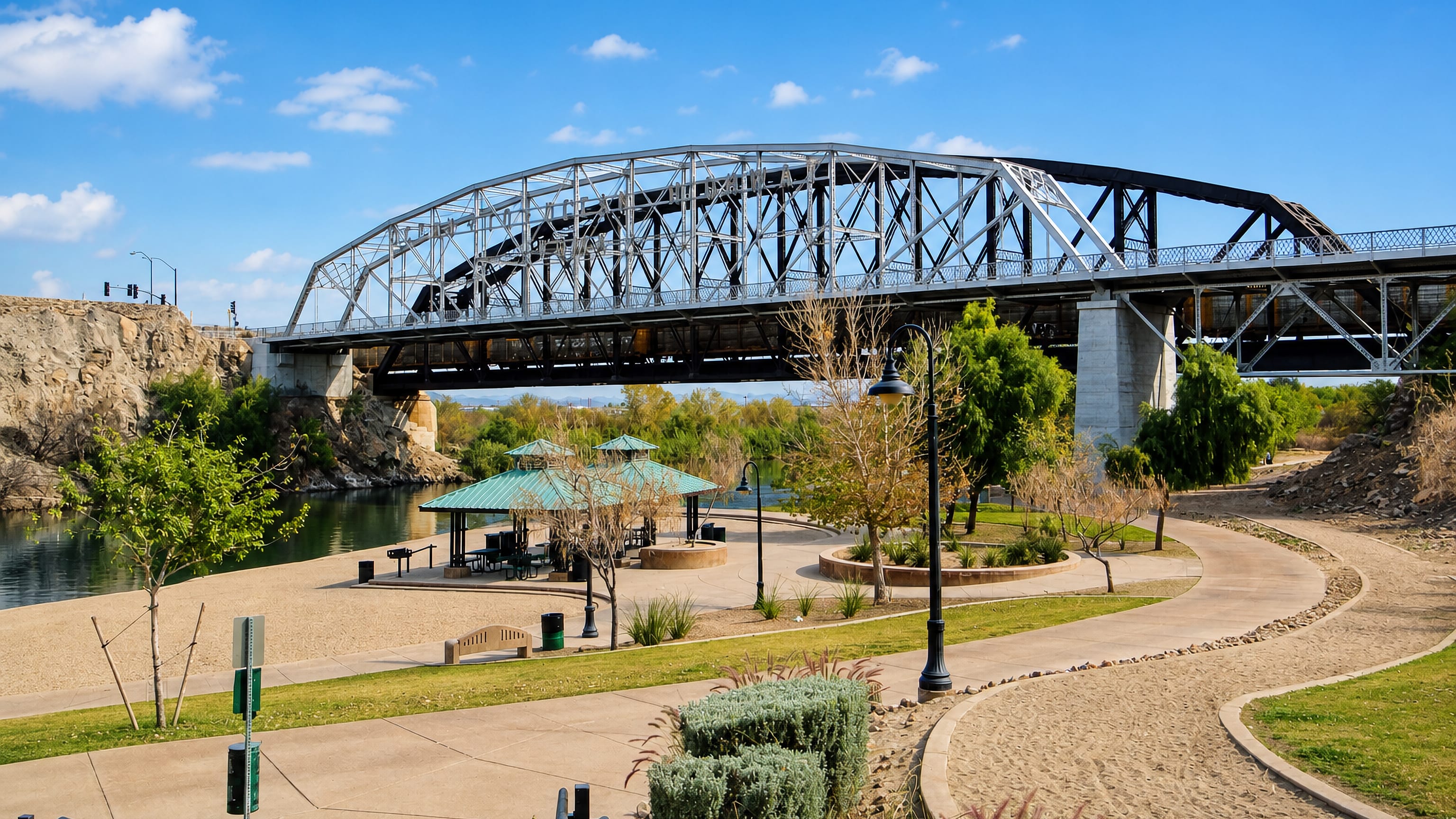 Steel truss bridge over the Colorado River at Yuma, Arizona, above a landscaped riverside park with shade ramadas.