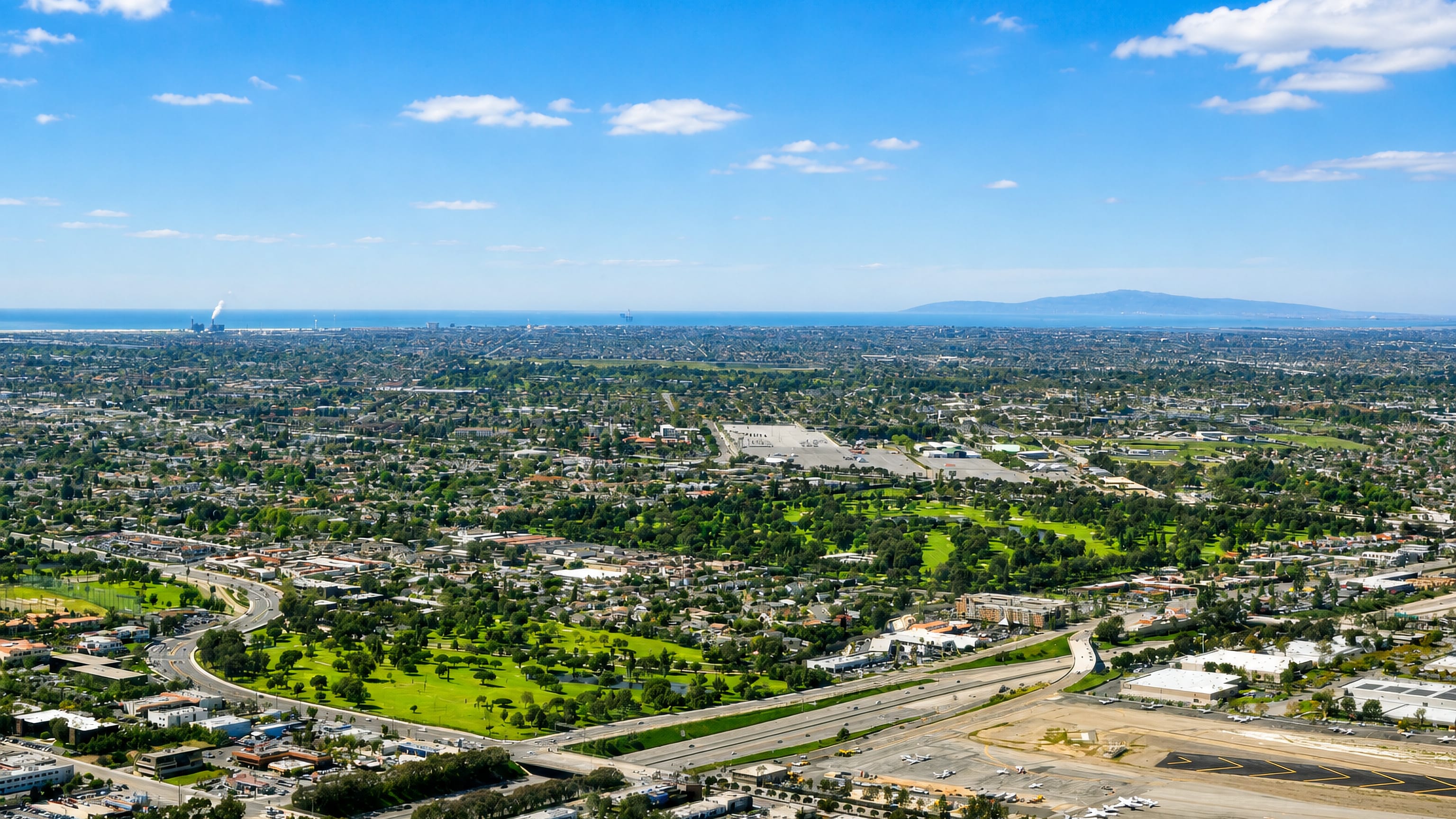 Aerial view over Costa Mesa, California, with a golf course, freeway, airport runway and the ocean beyond.