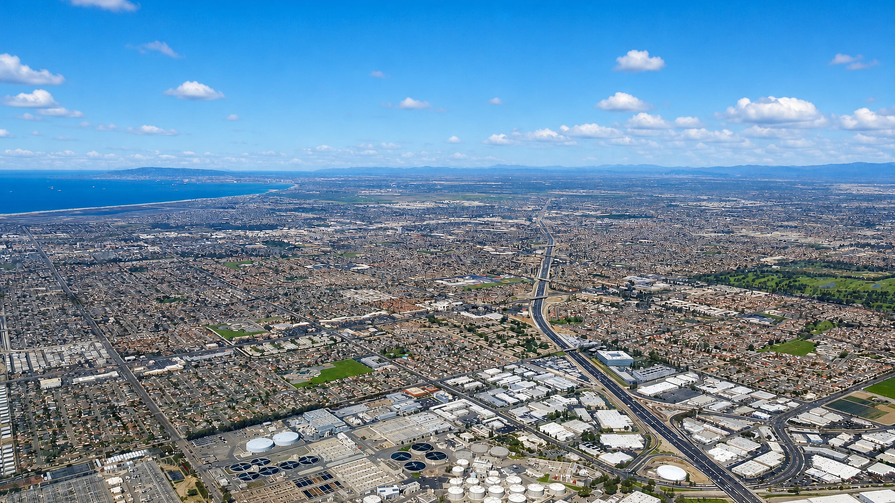 High aerial over Fountain Valley, California, its suburban grid reaching the coast past a treatment plant.