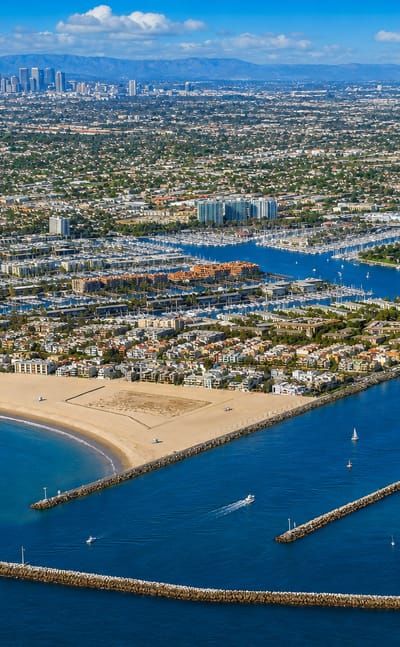 Aerial view of Marina del Rey harbor with moored boats, breakwaters, a sandy beach and the West LA skyline beyond