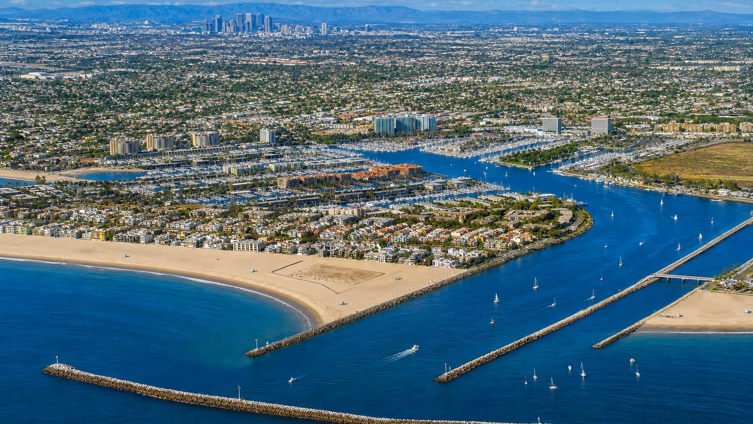 Aerial view of Marina del Rey harbor with moored boats, breakwaters, a sandy beach and the West LA skyline beyond