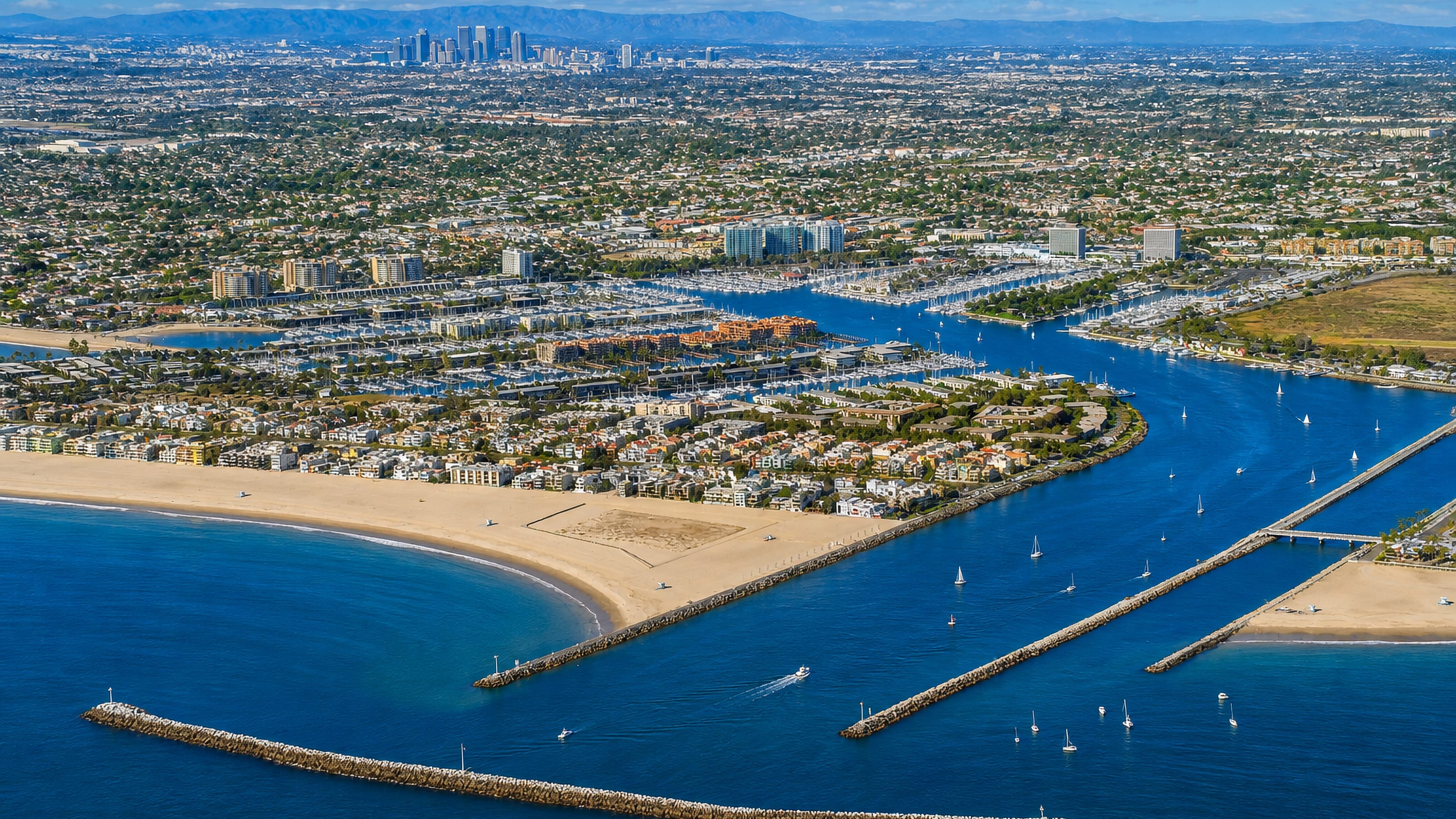 Aerial view of Marina del Rey harbor with moored boats, breakwaters, a sandy beach and the West LA skyline beyond