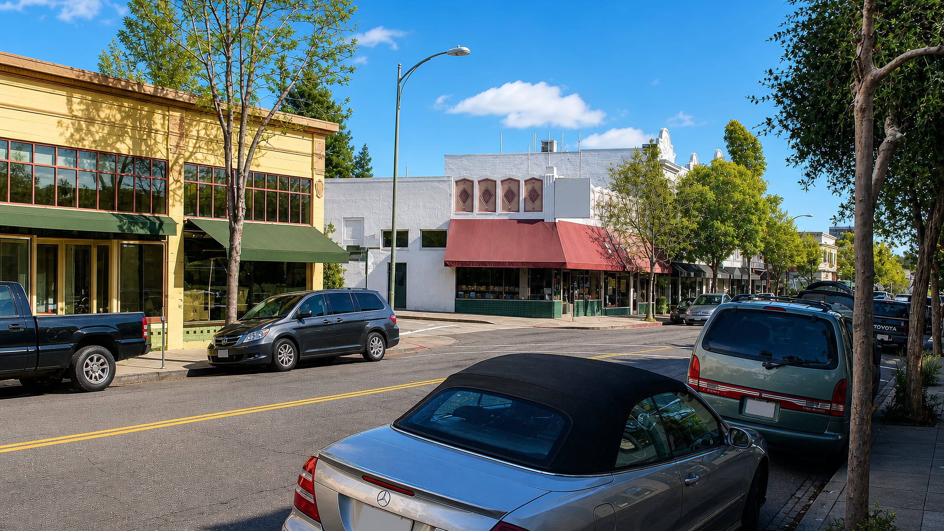 Low-rise shopfronts with awnings and parked cars along a sunlit commercial street in Rockridge, Oakland.