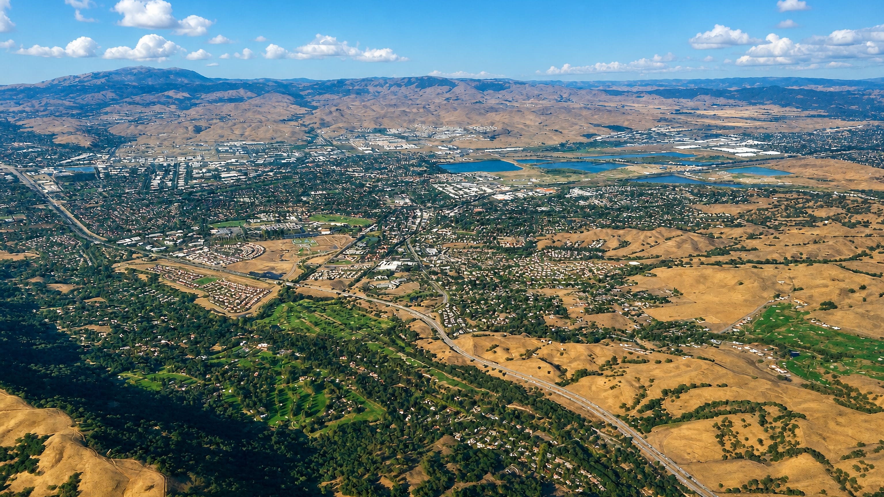 High aerial over Pleasanton, California, its neighbourhoods spread between golden hills and valley lakes.