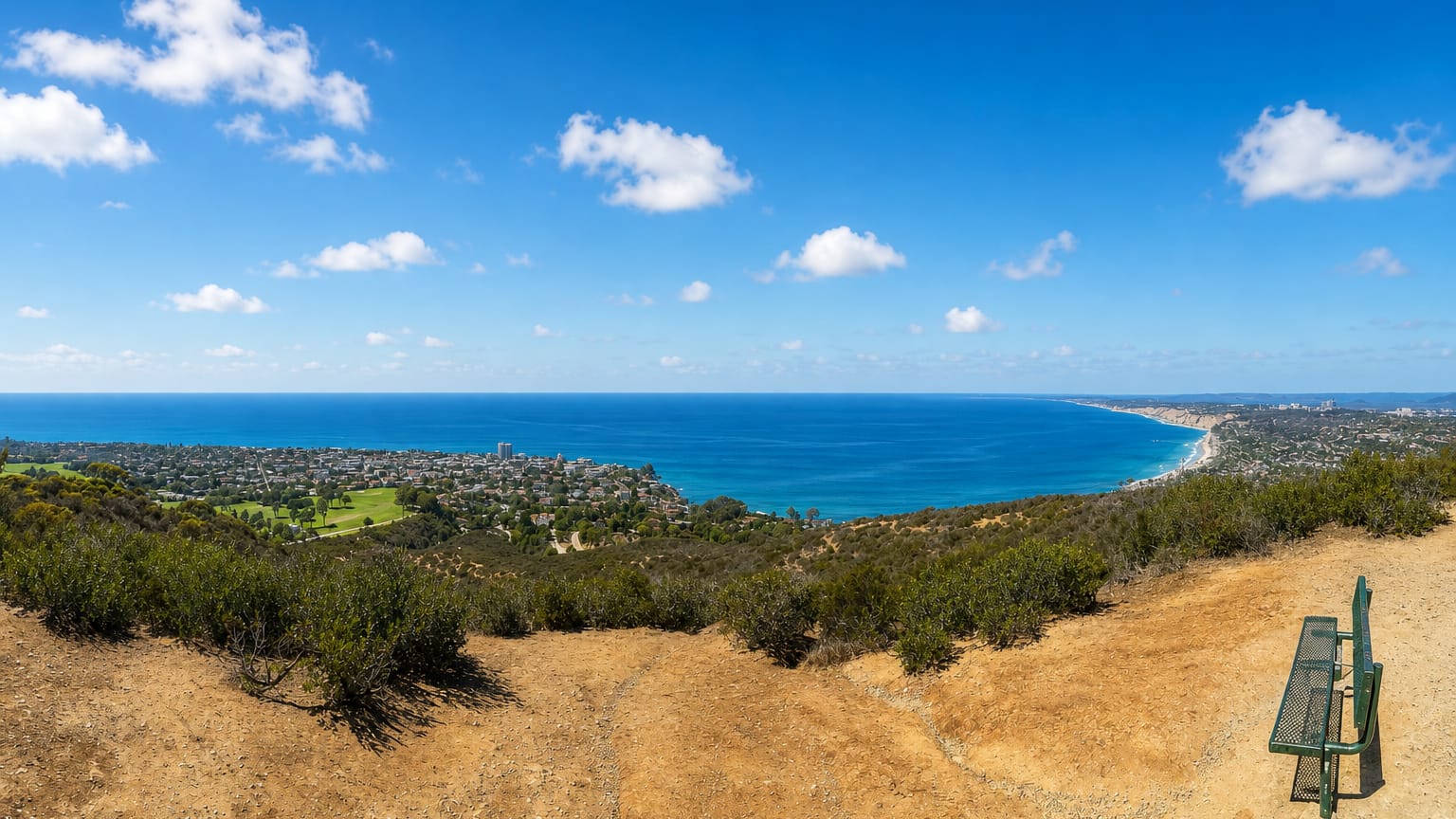 Panoramic coastal view over La Jolla and the Pacific Ocean from a hilltop trail with a lookout bench in San Diego.