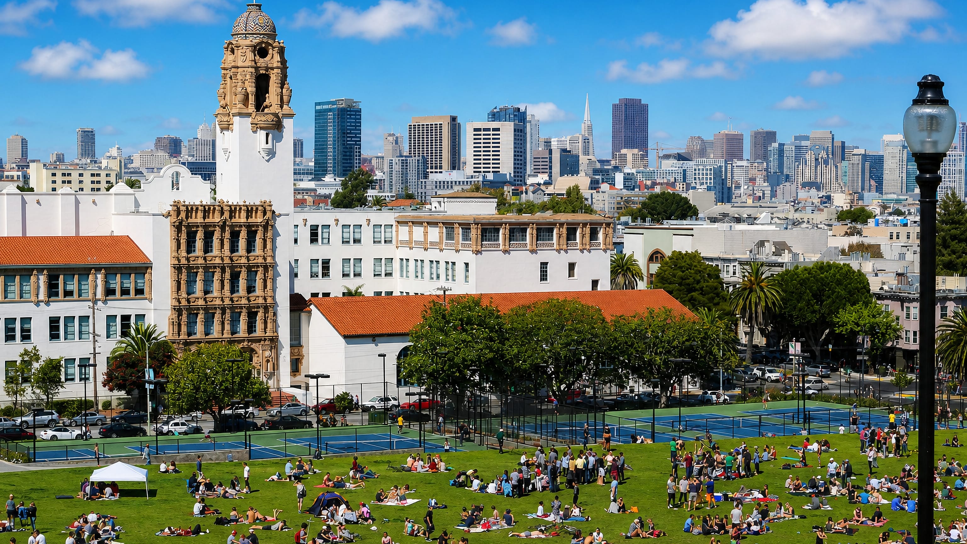 Crowds on the lawn at Dolores Park below Mission High School's tower and the San Francisco skyline