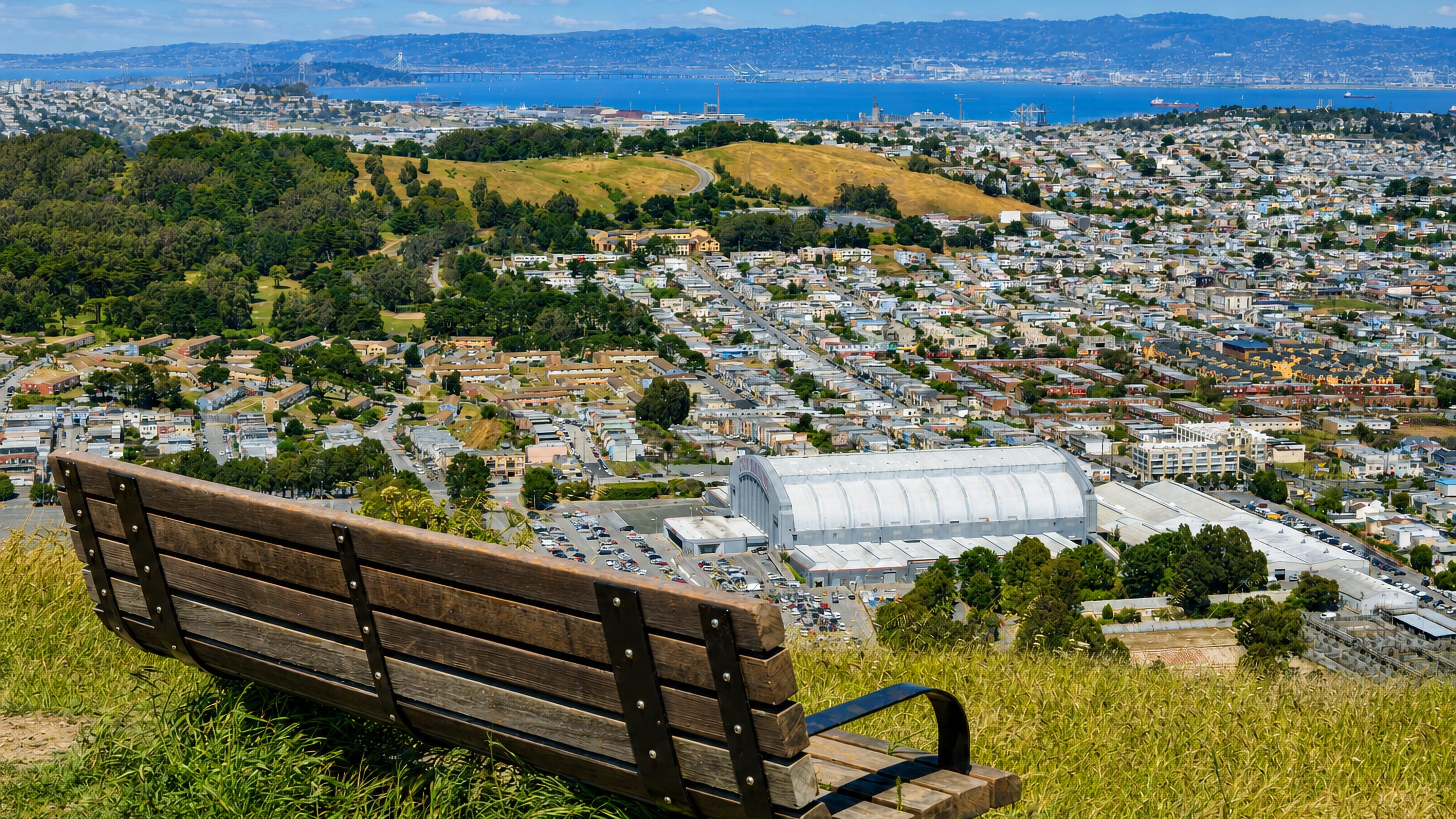 A hilltop bench overlooking Visitacion Valley in San Francisco, the arched Cow Palace roof and the bay beyond.