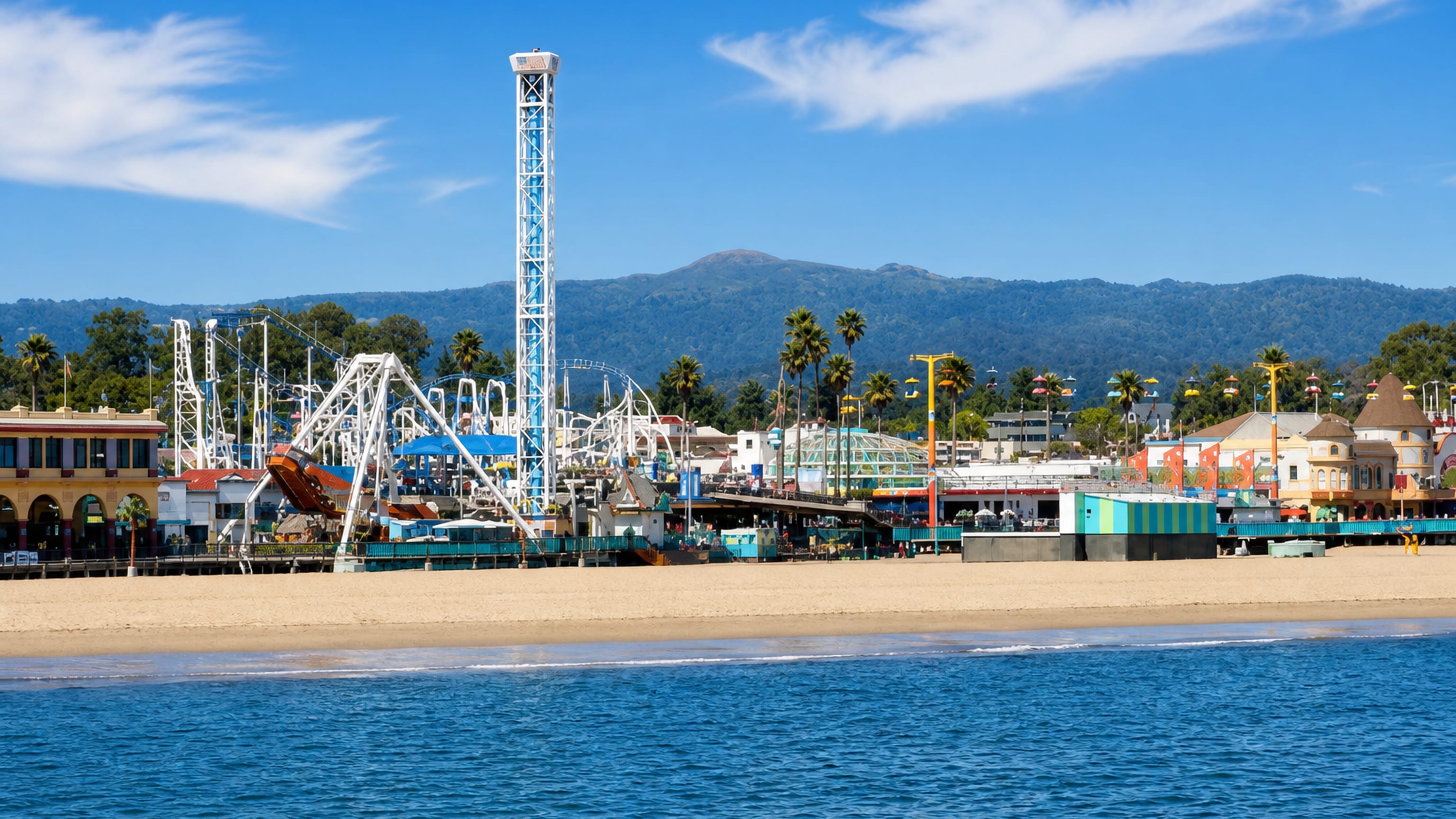Santa Cruz Beach Boardwalk with the Giant Dipper coaster and drop tower above the sand, mountains behind.