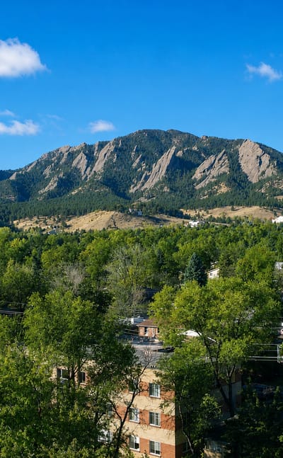 The Flatirons rock formations rising above green treetops and homes in Boulder, Colorado, on a clear day.