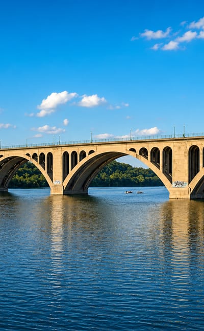 Arched Key Bridge spanning the Potomac River at Washington under a clear blue sky, with riverfront towers beyond.