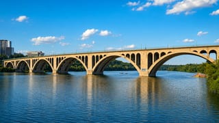 Arched Key Bridge spanning the Potomac River at Washington under a clear blue sky, with riverfront towers beyond.