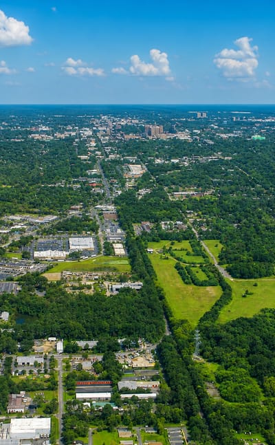 Aerial view over Gainesville, Florida's dense tree canopy toward the small distant downtown skyline under a blue sky.