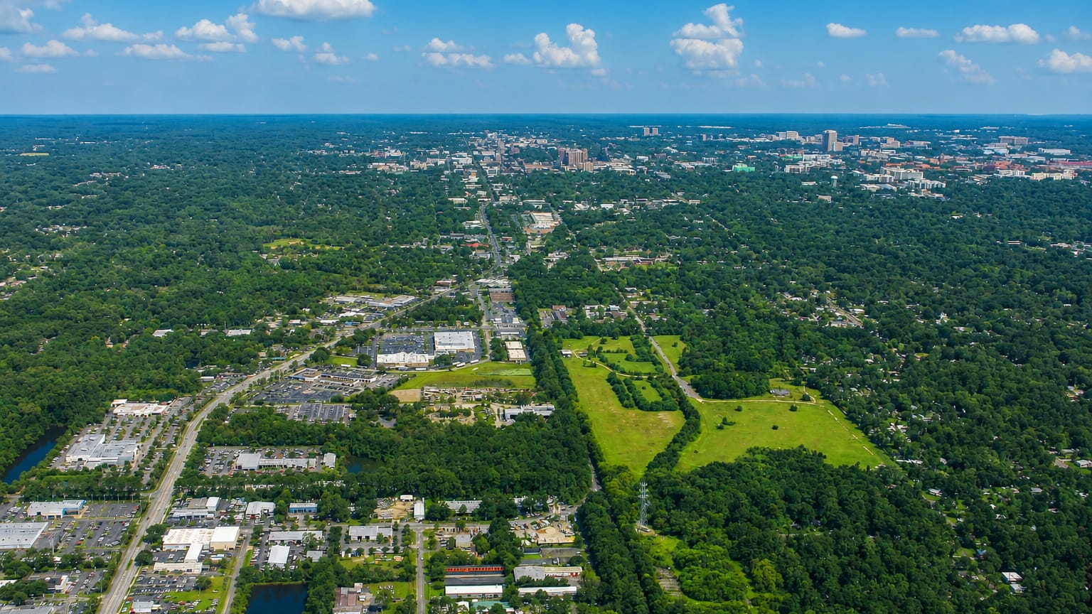 Aerial view over Gainesville, Florida's dense tree canopy toward the small distant downtown skyline under a blue sky.
