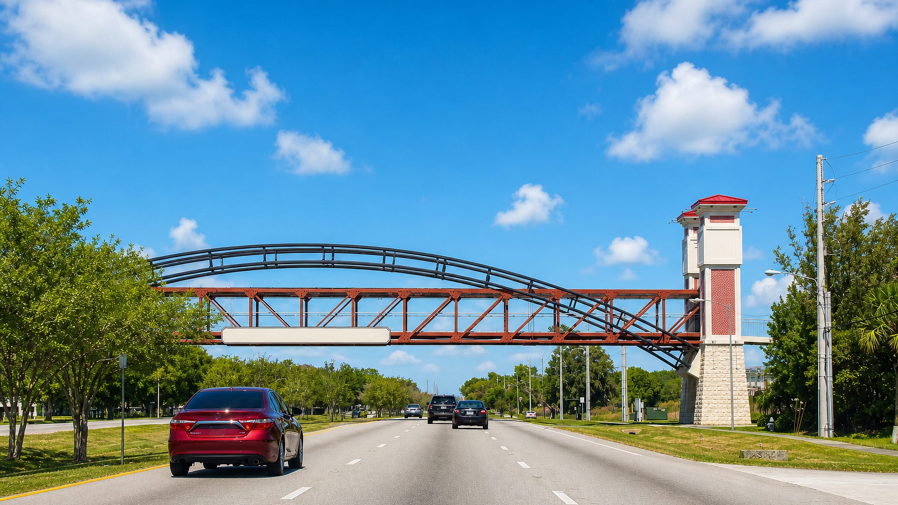 Divided highway in Kissimmee, Florida, passing beneath an arched pedestrian bridge under a bright blue sky.