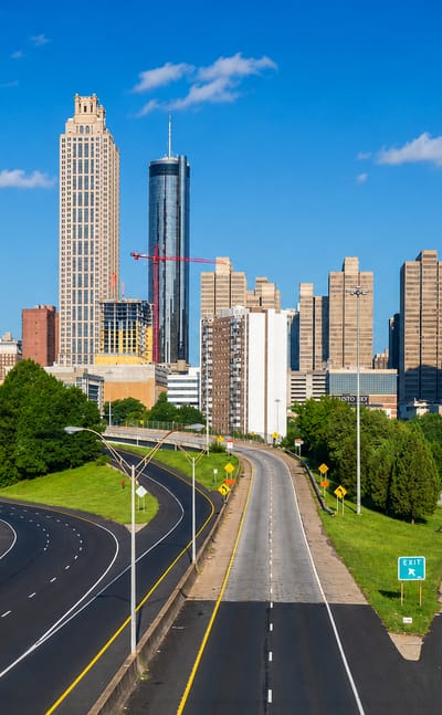 Downtown Atlanta skyline of tall towers viewed from a curving highway lined with green trees under blue sky
