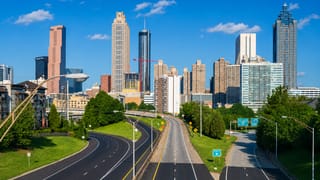 Downtown Atlanta skyline of tall towers viewed from a curving highway lined with green trees under blue sky