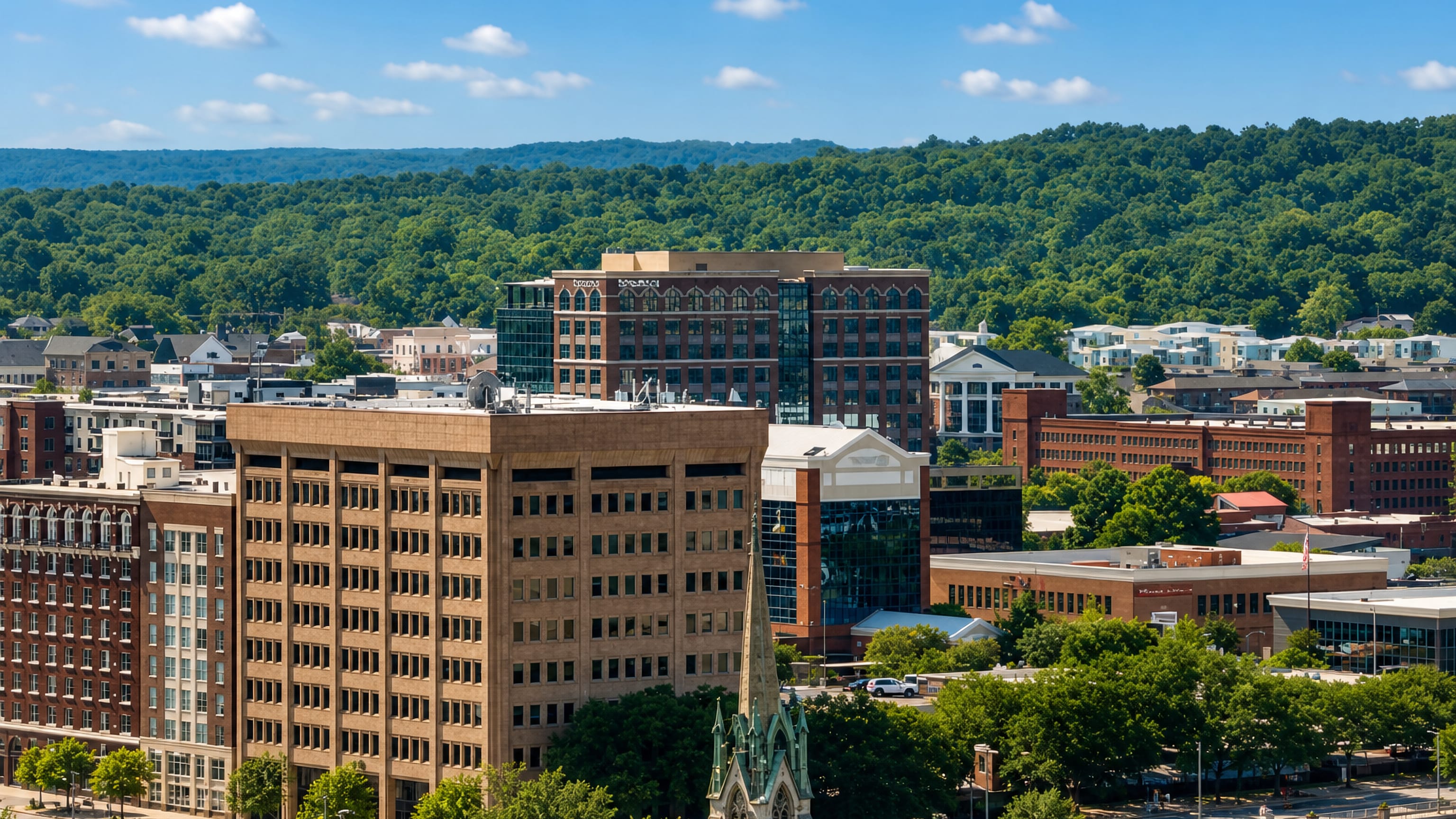Downtown Columbus, Georgia, with riverfront office towers, brick mill buildings and a church spire below wooded hills