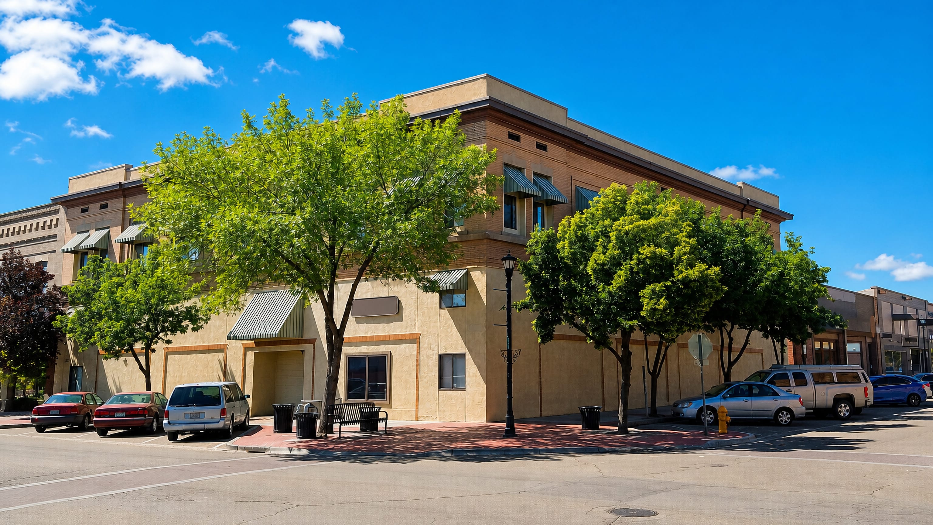 A tree-lined downtown street corner with low brick and stucco storefronts in Nampa, Idaho.