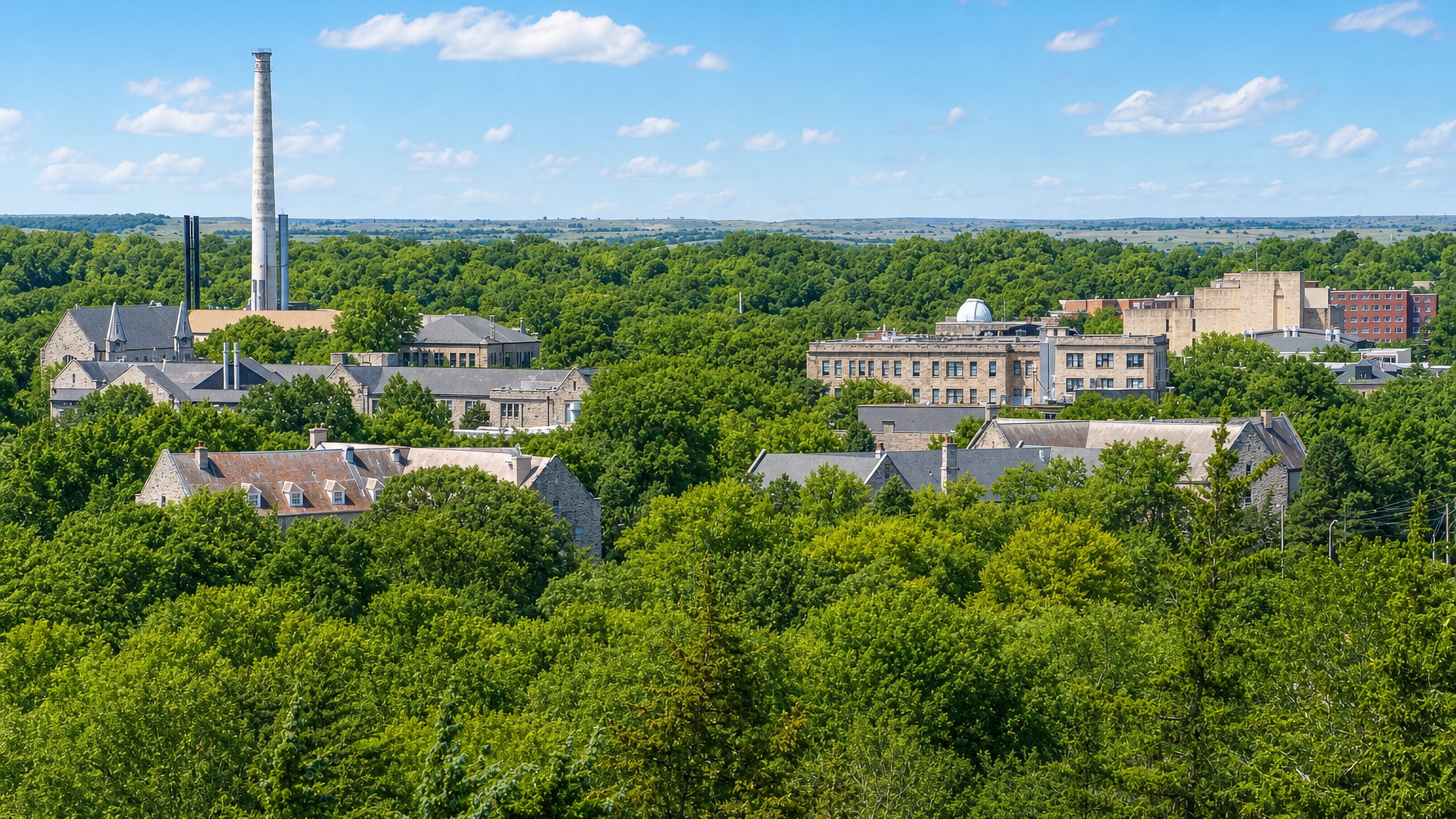 Limestone Kansas State University buildings among summer trees in Manhattan, Kansas, with Flint Hills beyond.