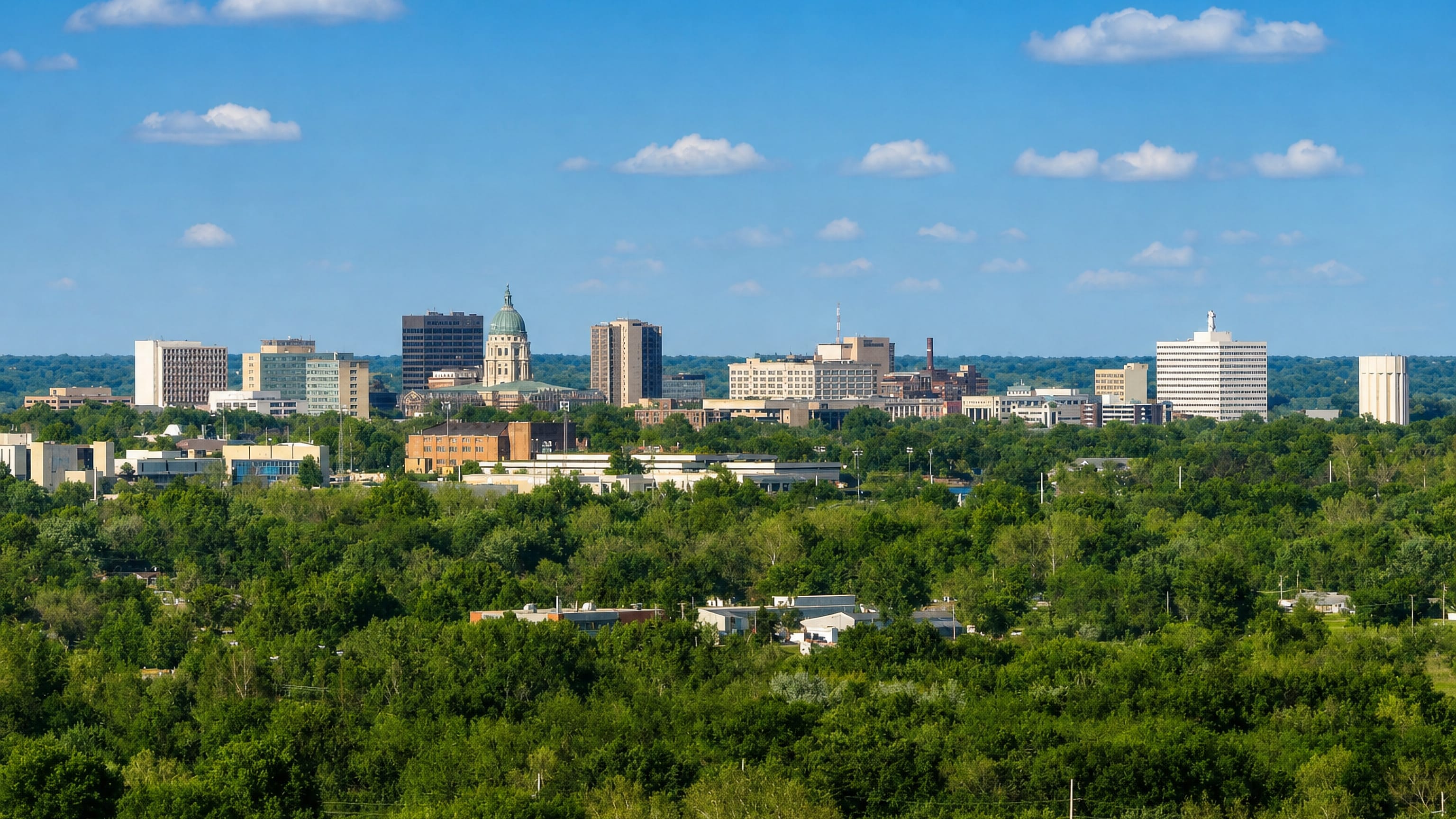 Topeka's skyline over wooded Kansas countryside, centred on the green copper dome of the Kansas State Capitol.