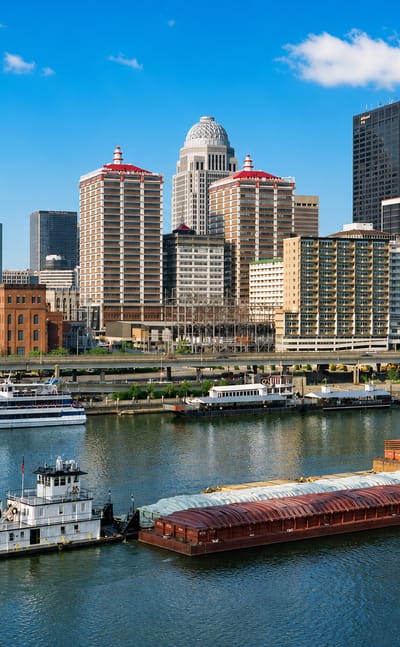 Louisville skyline along the Ohio River with a towboat pushing barges past the domed downtown buildings.