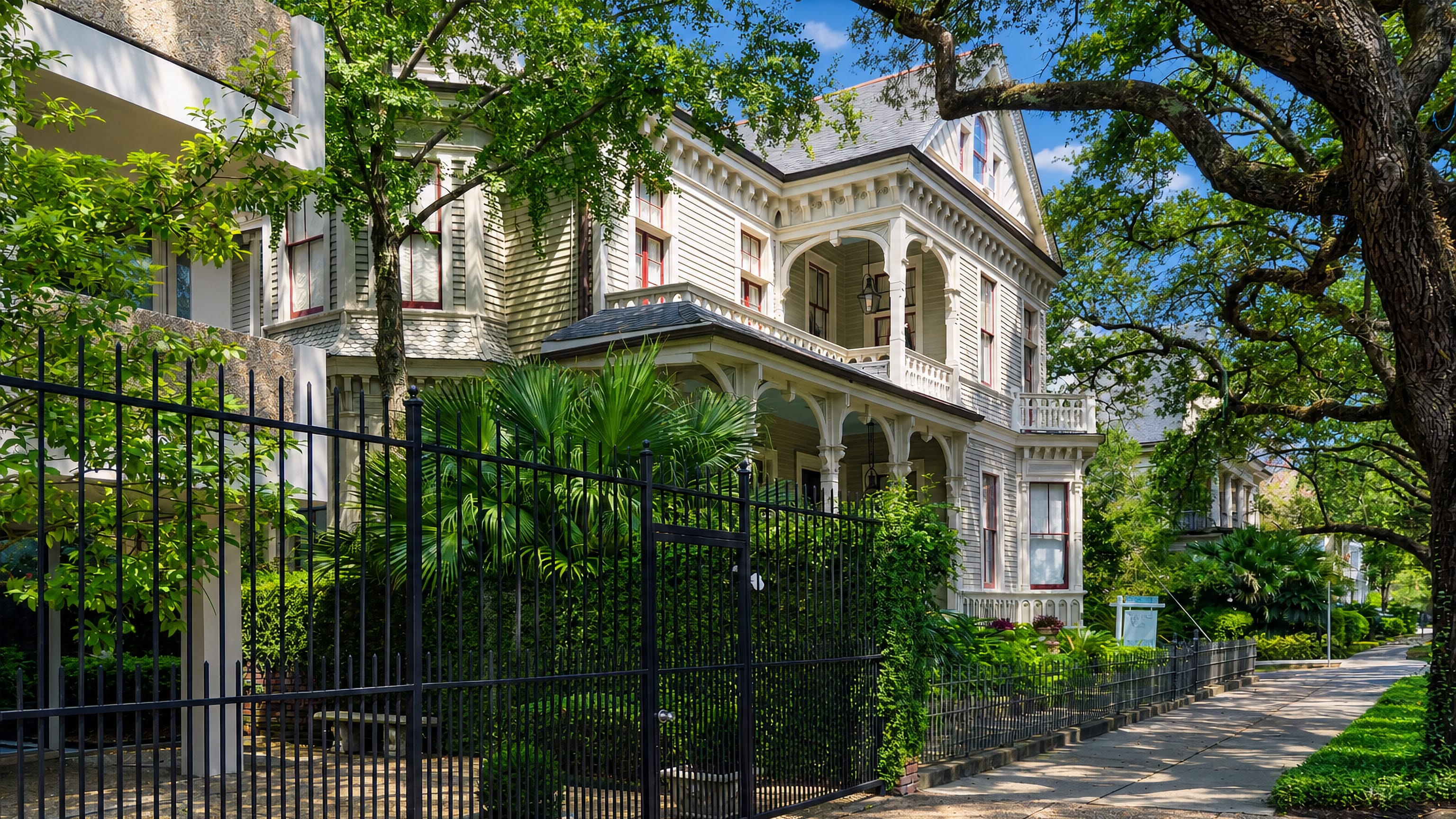 A galleried Victorian mansion behind iron railings under live oaks in New Orleans' Garden District.