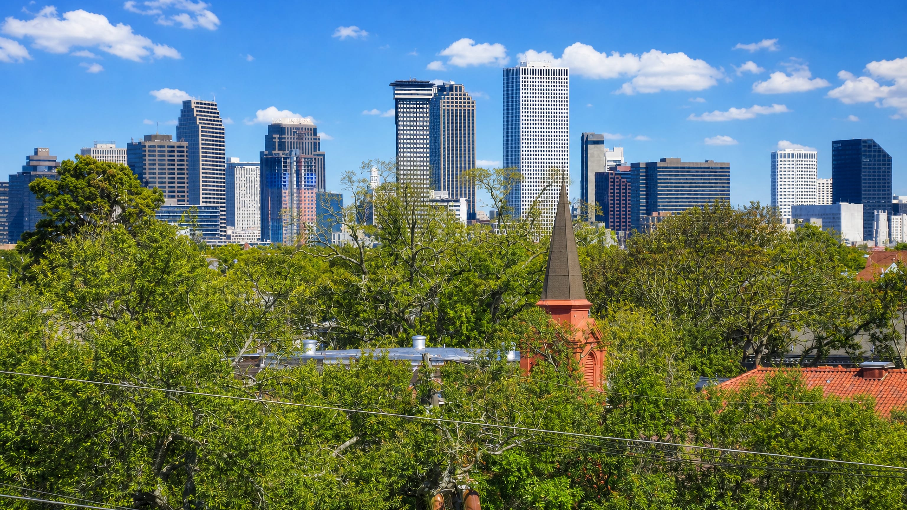 New Orleans downtown skyline rising beyond the oak canopy and a red-brick church steeple in Uptown