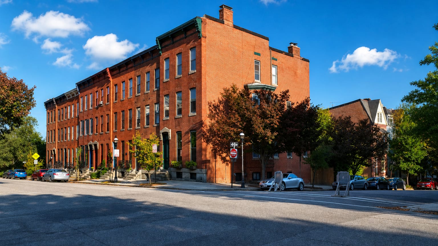 A sunlit block of three-story red-brick rowhouses on a quiet street corner in Baltimore's Bolton Hill.