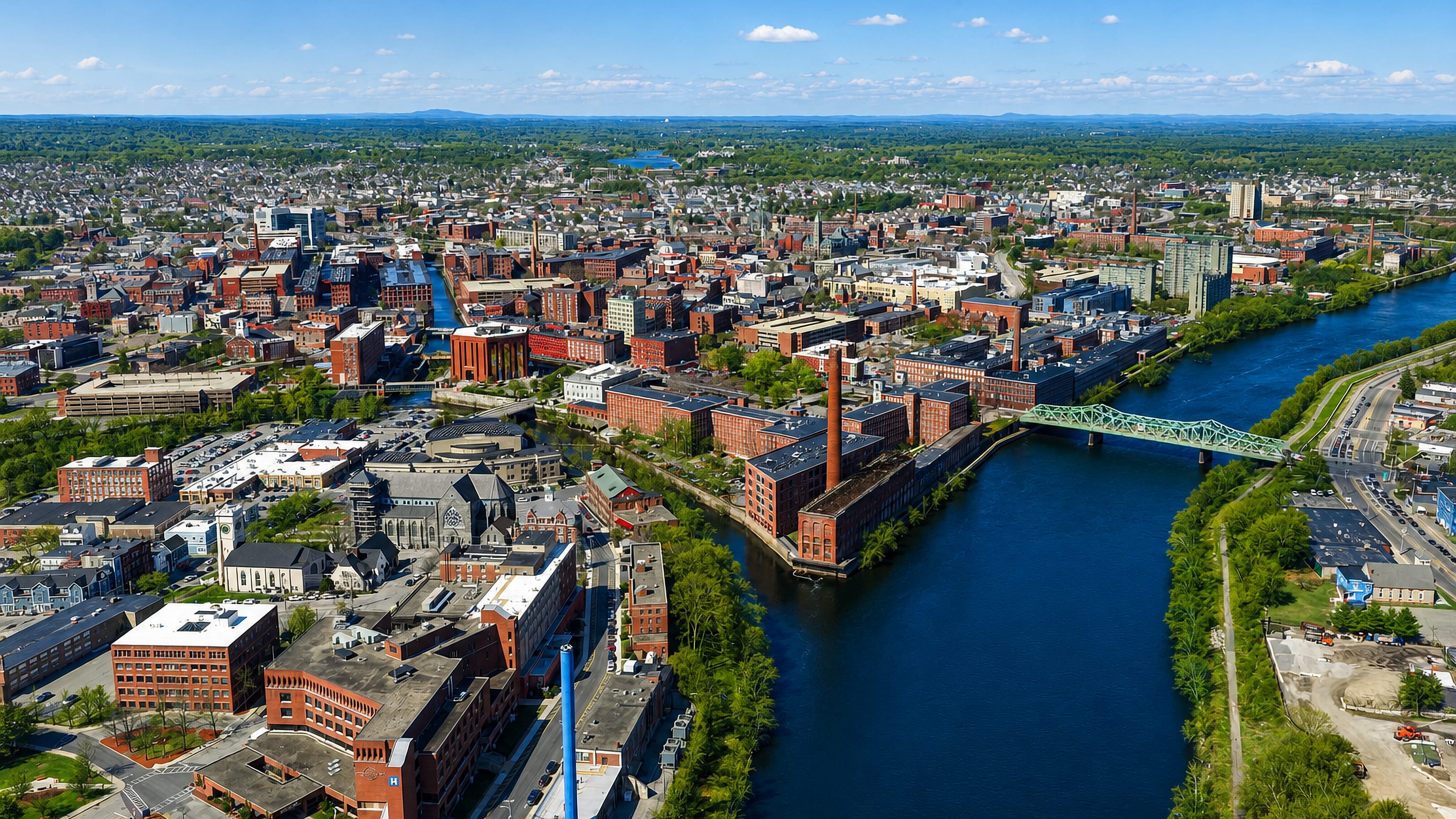 Aerial view of Lowell's red-brick textile mills and canals along the Merrimack River, crossed by a green truss bridge.