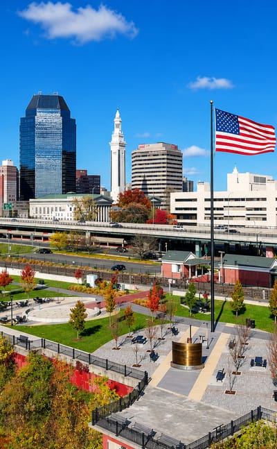 Springfield, Massachusetts, skyline with the Campanile and glass towers seen from a riverfront park flying a US flag.