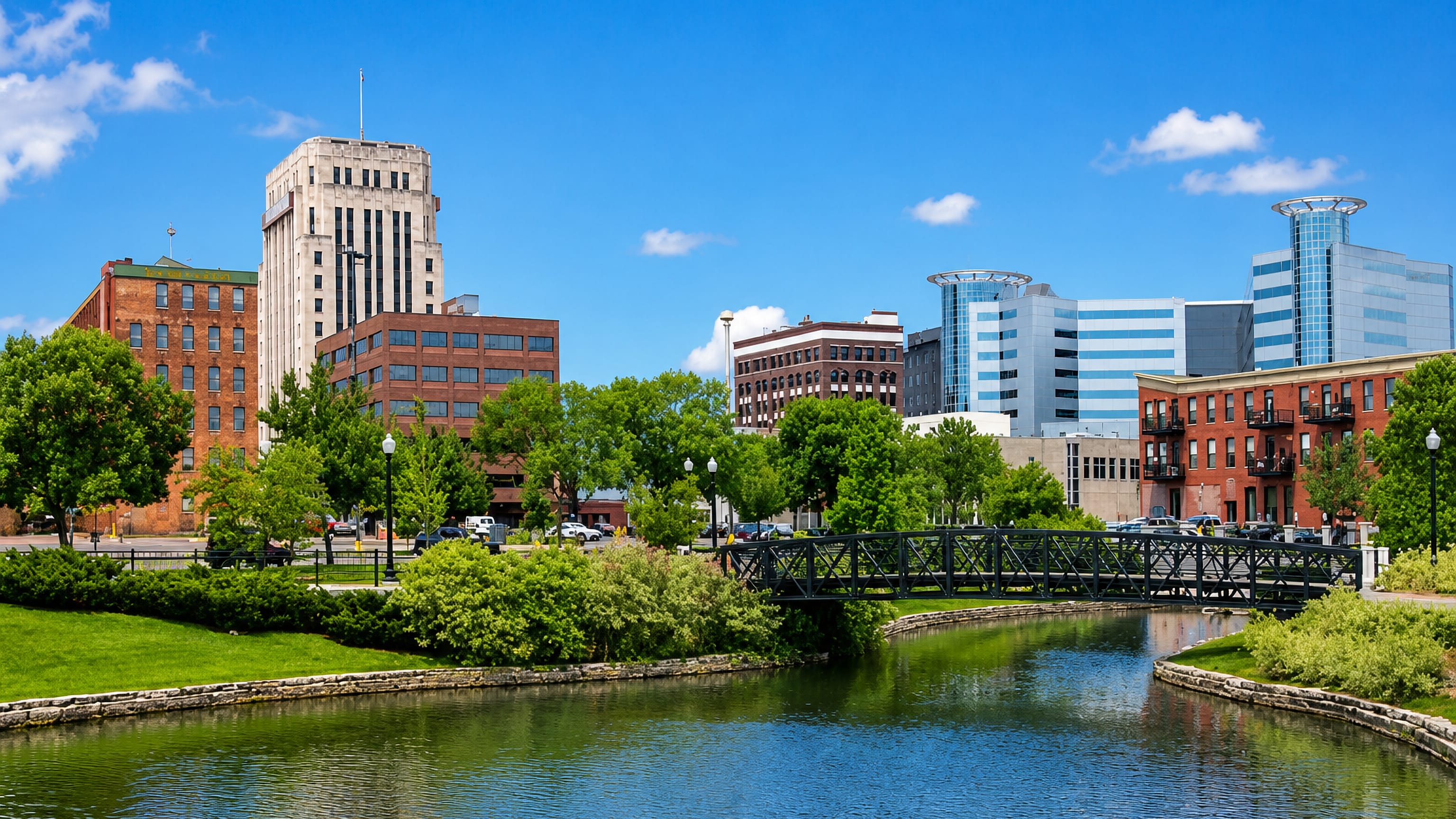 Downtown Kalamazoo's Art Deco tower and blue glass office blocks behind an arched footbridge over a tree-lined pond
