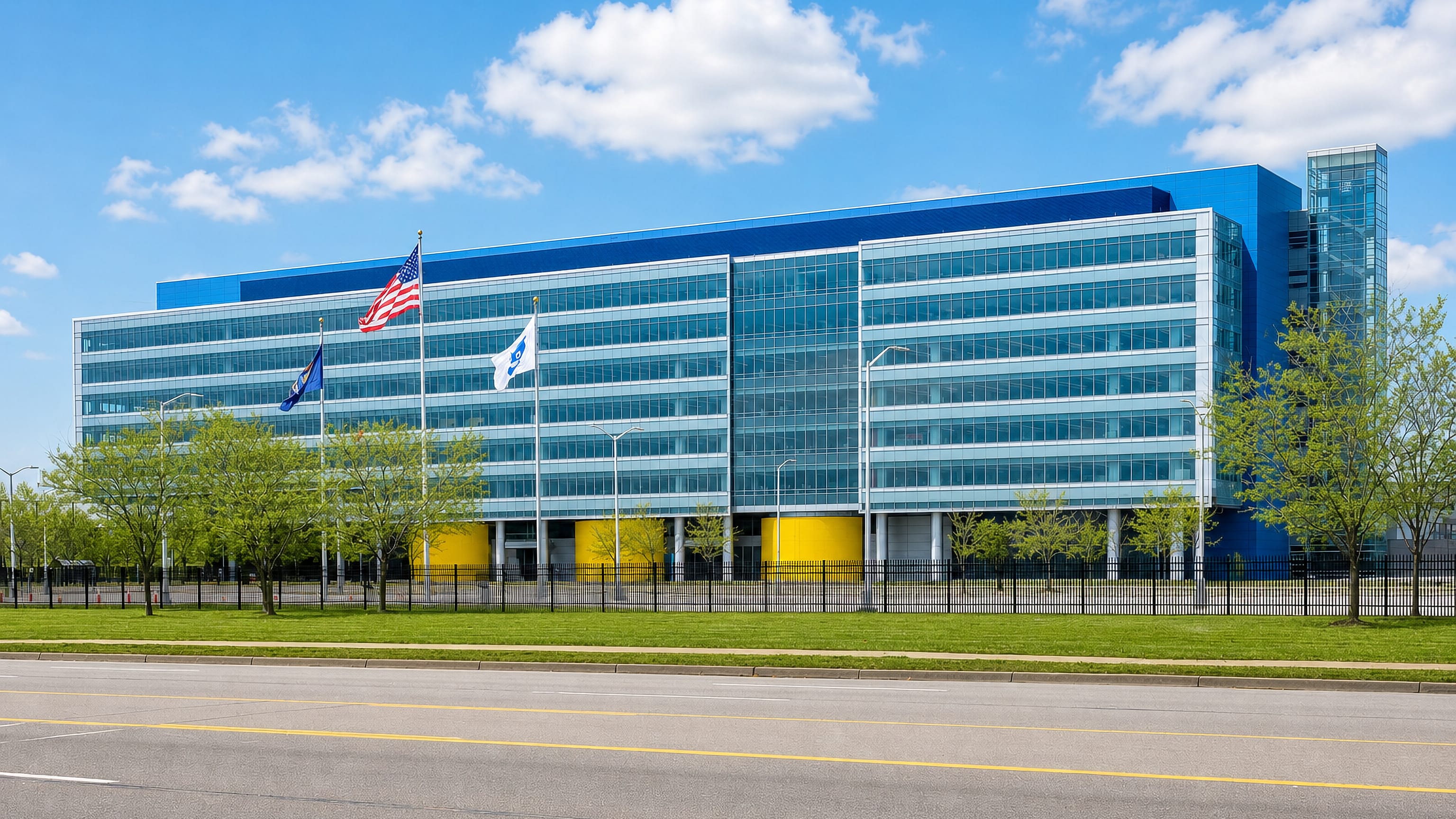 A modern blue-and-glass corporate office building behind flagpoles and lawn in Warren, Michigan.