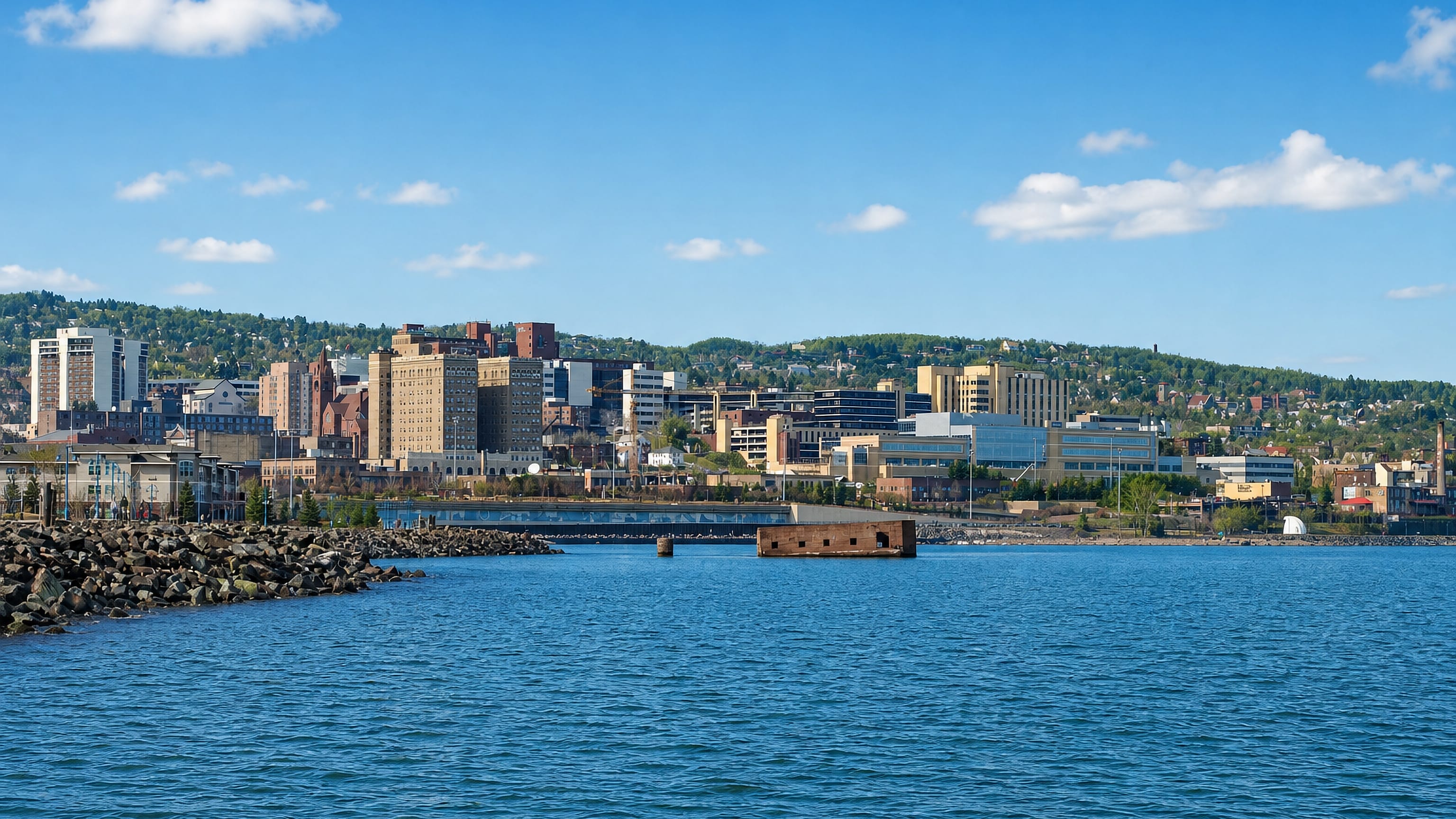 Duluth, Minnesota's hillside downtown rising above Lake Superior, seen past a rocky breakwater.