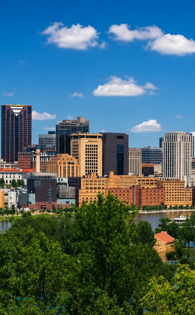 Saint Paul, Minnesota downtown skyline across the Mississippi River, framed by green riverbank trees.