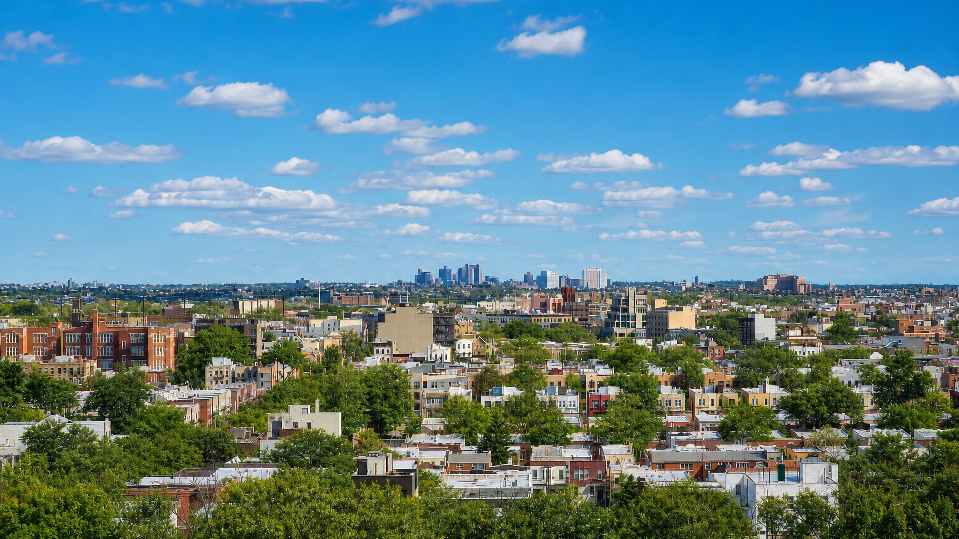 Low-rise rooftops and leafy streets of Astoria, Queens, with a distant cluster of towers on the horizon.
