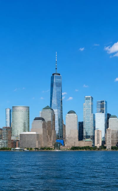 Lower Manhattan skyline across the Hudson River, dominated by One World Trade Center under a blue sky.