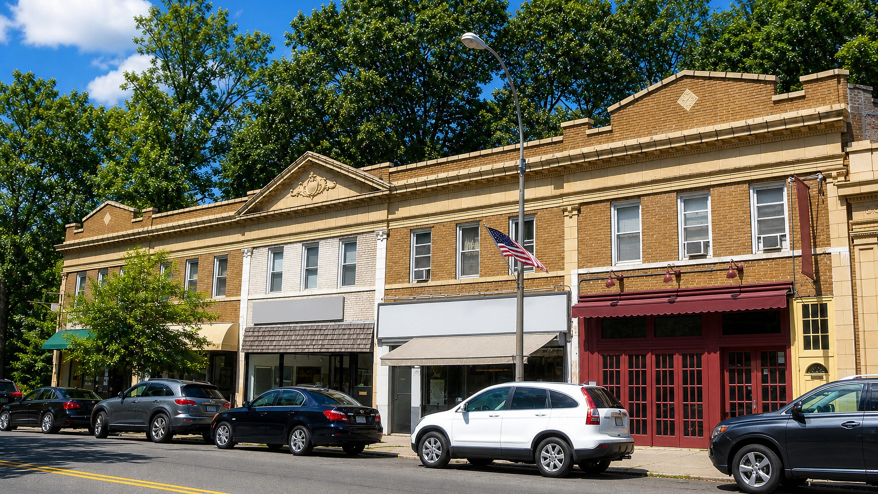 Early-20th-century brick shopfront row and parked cars along Douglaston's main street in Queens.