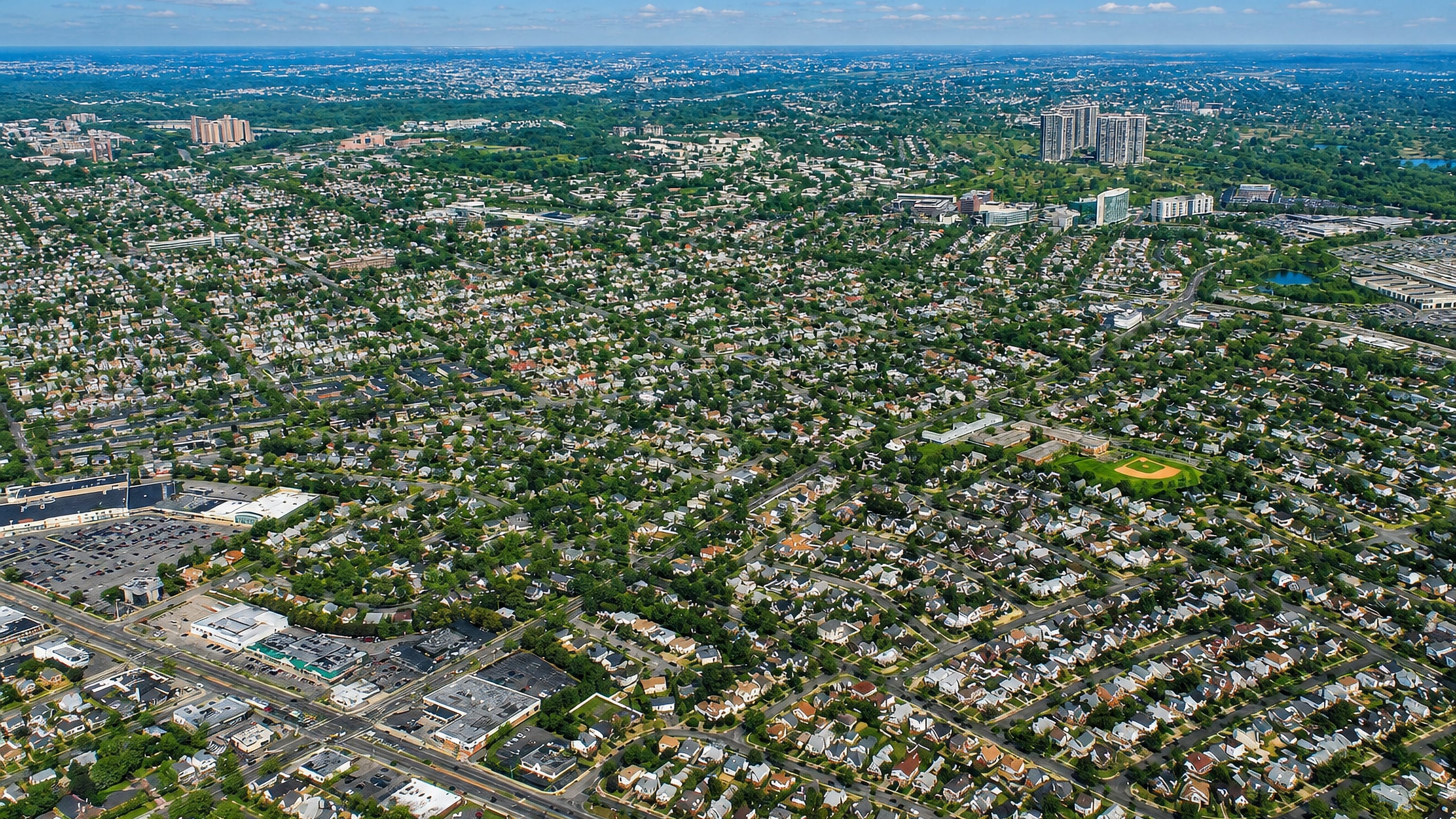 Aerial sweep of tree-lined single-family streets and a tower cluster in Glen Oaks, Queens, New York City.