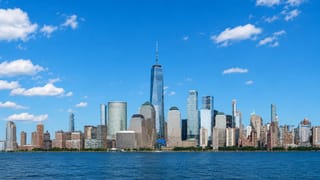 Lower Manhattan skyline across the Hudson River, dominated by One World Trade Center under a blue sky.