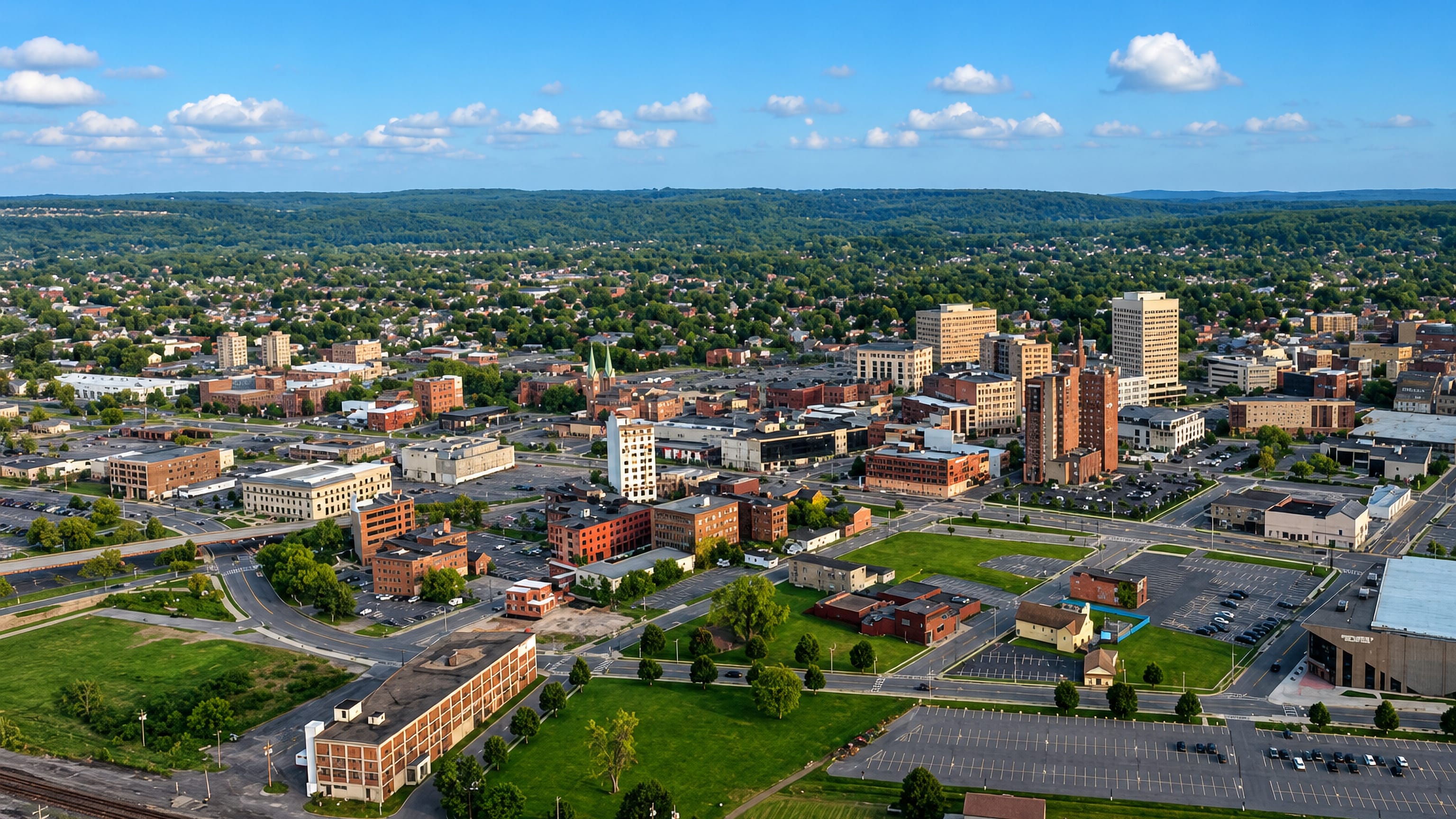 Aerial view of downtown Utica, New York, its brick towers and parking lots against the wooded Mohawk Valley hills.