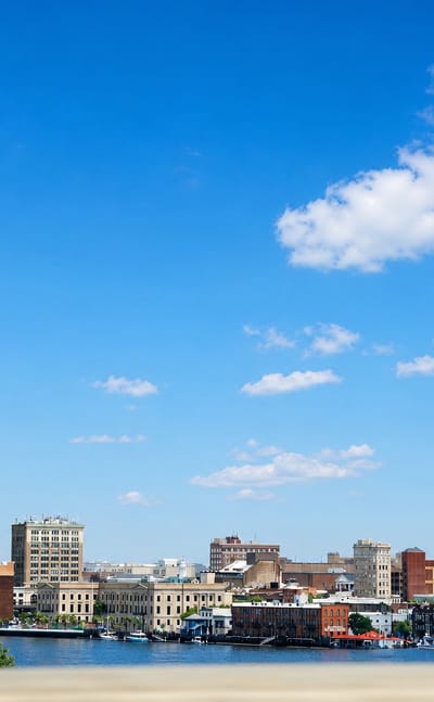 Downtown Wilmington, North Carolina's low riverfront skyline across the Cape Fear River under a wide blue sky.