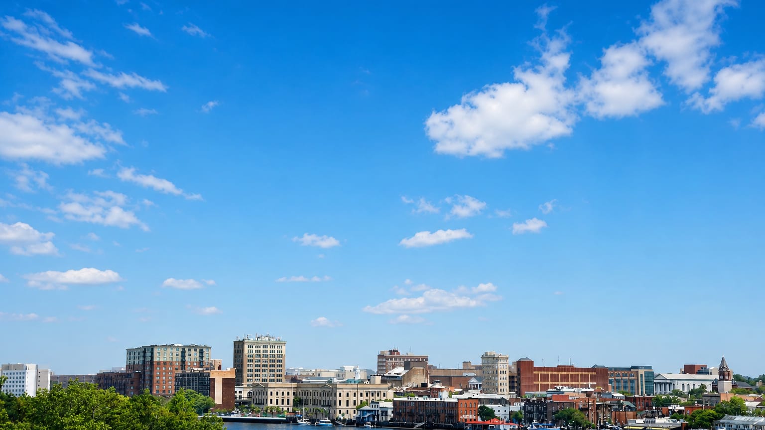 Downtown Wilmington, North Carolina's low riverfront skyline across the Cape Fear River under a wide blue sky.