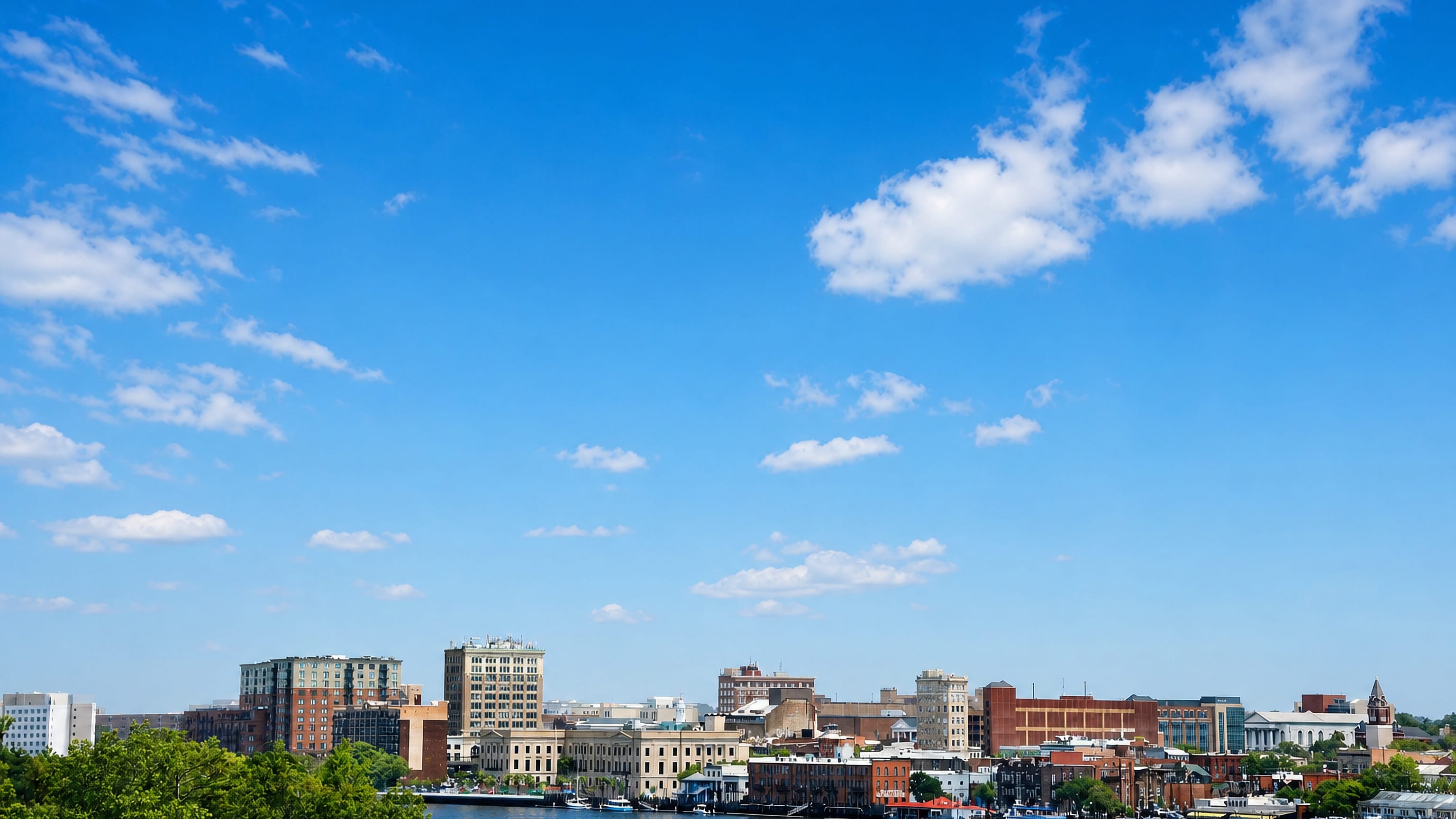 Downtown Wilmington, North Carolina's low riverfront skyline across the Cape Fear River under a wide blue sky.
